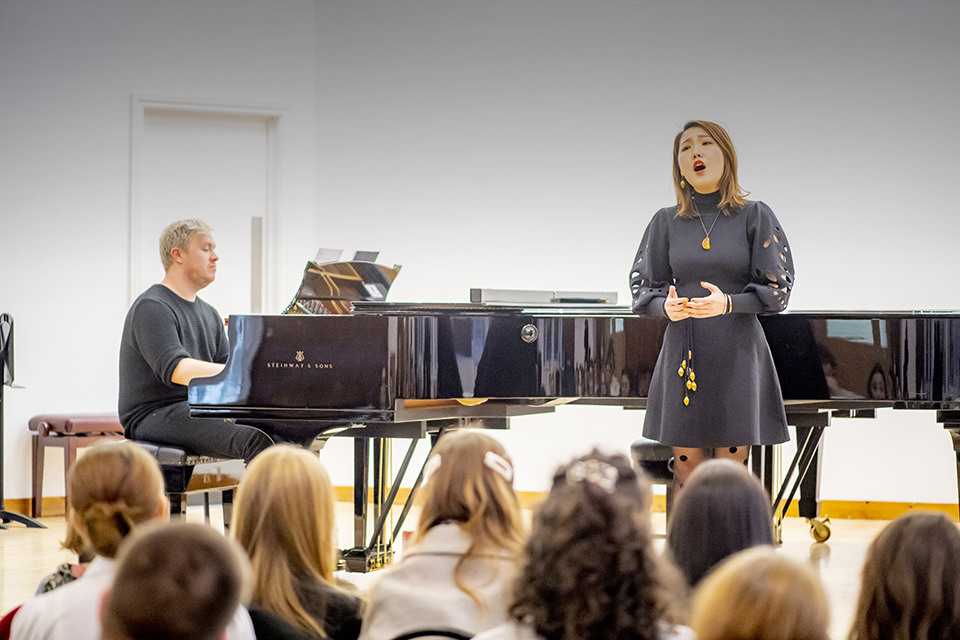 A female student singing on stage with piano accompaniment to a room of students.